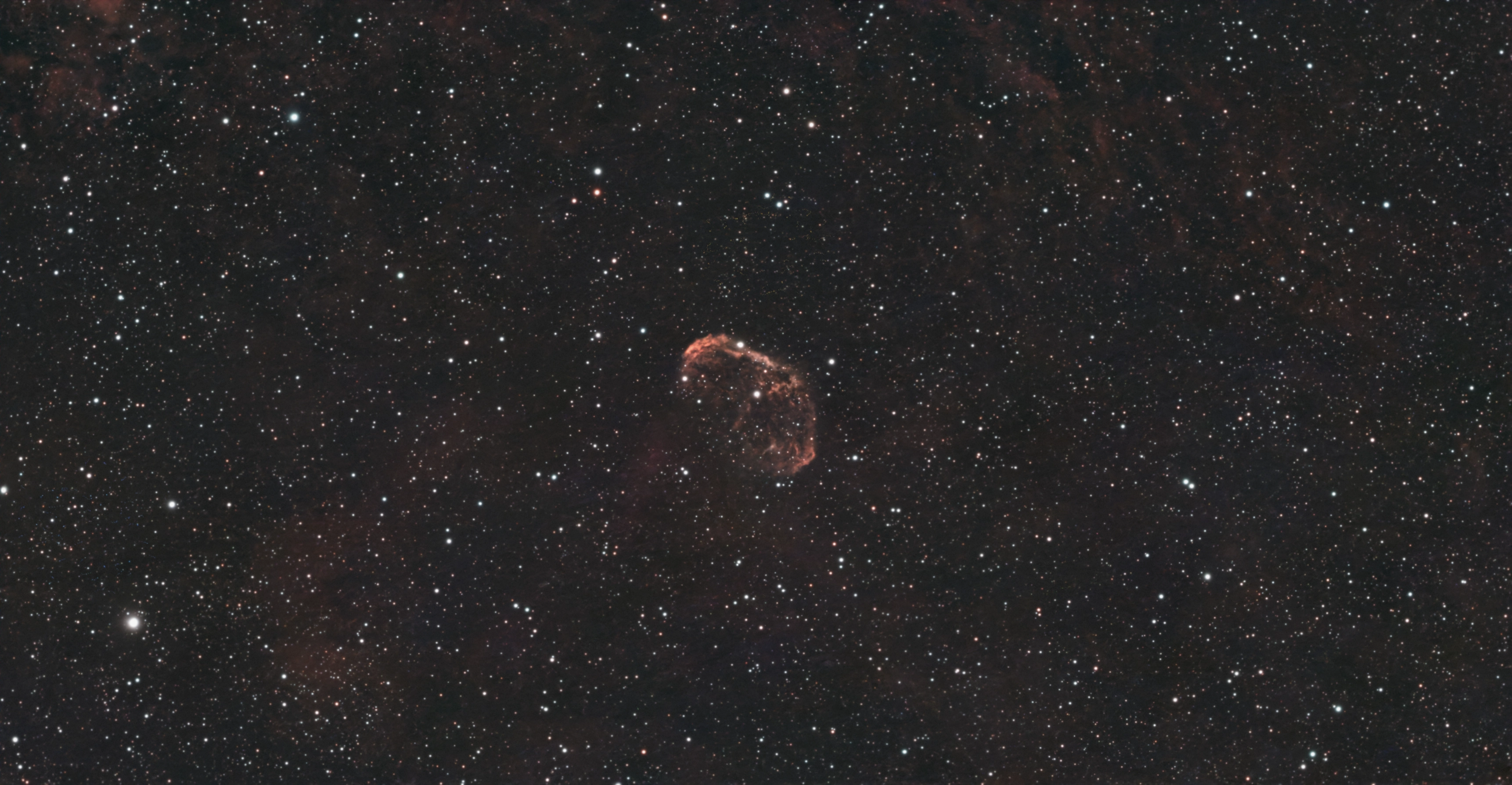 Curved red shell of the Crescent Nebula amid faint red gas and a dense star field